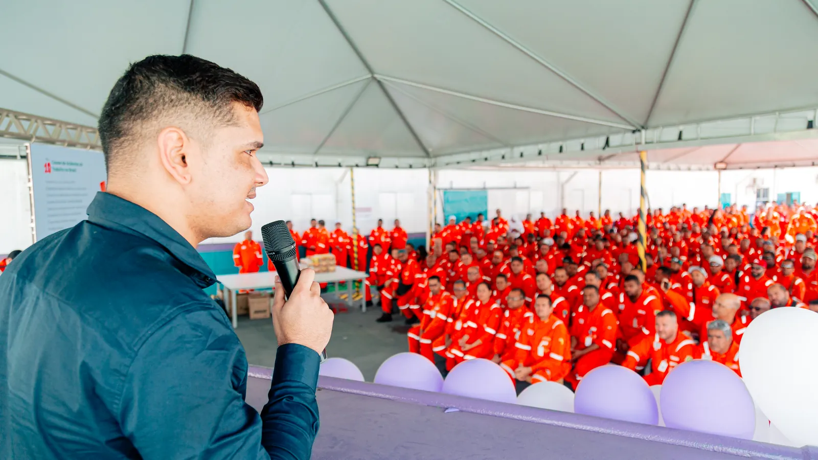 Deivson Lest ao microfone diante de centenas de trabalhadores de uniforme laranja sob uma tenda.